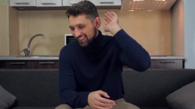 Koyu mavi kazaklı ve gri pantolonlu genç bir adam kanepede oturuyor, TV 'de eğlence izliyor, parmağını gösterip gülüyor. Mutfak arka planda. 4k yavaş çekim görüntüsü