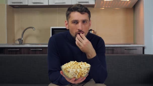 一个身穿深蓝色套头毛衣和灰色裤子的年轻人坐在沙发上 用透明的玻璃盘子吃爆米花 并在电视上观看娱乐节目 厨房的背景 4K慢镜头 — 图库视频影像