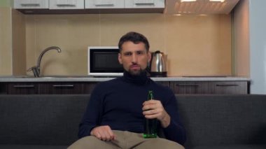 Koyu mavi kazaklı ve gri pantolonlu ciddi bir genç adam kanepede oturuyor, bira içiyor ve TV 'de eğlence izliyor. Mutfak arka planda. 4k yavaş çekim görüntüsü