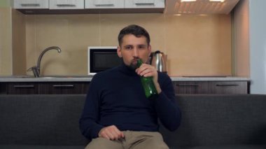 Koyu mavi kazaklı ve gri pantolonlu ciddi bir genç adam kanepede oturuyor, bira içiyor ve TV 'de eğlence izliyor. Mutfak arka planda. 4k yavaş çekim görüntüsü