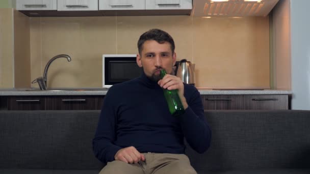 严肃的年轻人穿着深蓝色套头毛衣和灰色裤子坐在沙发上 喝啤酒 看电视上的娱乐节目 厨房的背景 4K慢镜头 — 图库视频影像