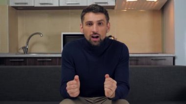 Koyu mavi kazaklı ve gri pantolonlu genç bir adam TV 'de spor maçı izliyor, kanepeden kalkın ve favori takımının zaferinin tadını çıkarın. Mutfak arka planda. 4k yavaş çekim görüntüsü