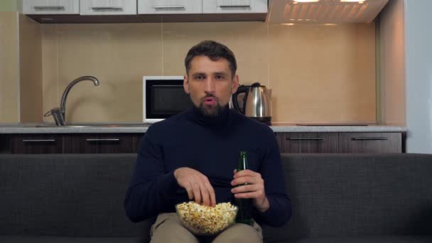 ソファに座っている濃い青のプルオーバーとグレーのパンツの若い男は ポップコーンを食べ ビールを飲み テレビでエンターテイメントを見る キッチンを背景に 4Kスローモーション映像 — ストック動画