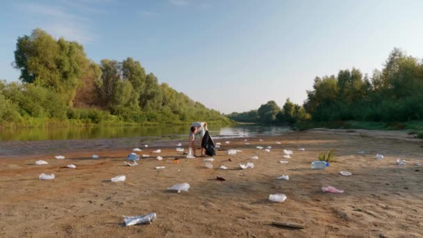 Um homem é coleta lixo de plástico nas margens de um rio ou lago seco e poluído — Vídeo de Stock