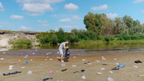 男は、乾燥した汚染された川や湖のほとりにプラスチックごみを収集しています — ストック動画