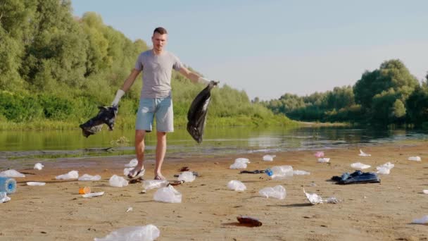 Adam plastik bir torba yırtıyor ve kirlenmiş nehrin kıyısında çöplerin arasında yürüyor. — Stok video