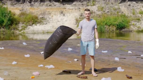 Man is stands with plastic bag along the trash on the banks of polluted river — 비디오