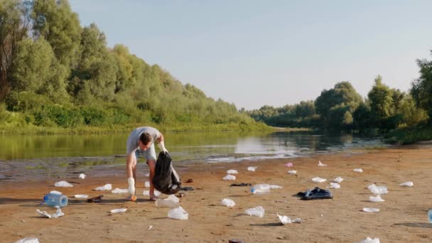 男は汚染された川のほとりにプラスチックごみを収集し、親指を表示されます. — ストック動画