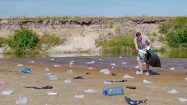 Hombre bailando y recoge basura plástica en las orillas del río seco y contaminado — Vídeos de Stock