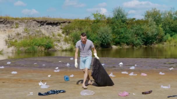De mens verzamelt plastic afval op de oevers van vervuilde rivieren en luistert naar muziek. — Stockvideo