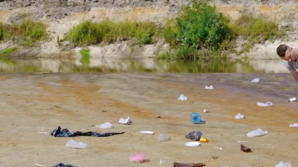 Människan samlar plast skräp på stranden av förorenade floden och lyssnar på musik — Stockvideo