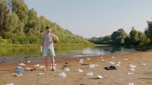Mannen plockar upp plastskräp på stranden av förorenade floden och visar ogillande. — Stockvideo