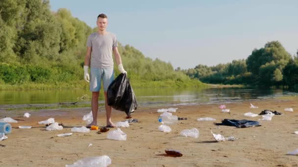 Man torkar svett från ansiktet bland soporna på stranden av förorenad flod — Stockvideo