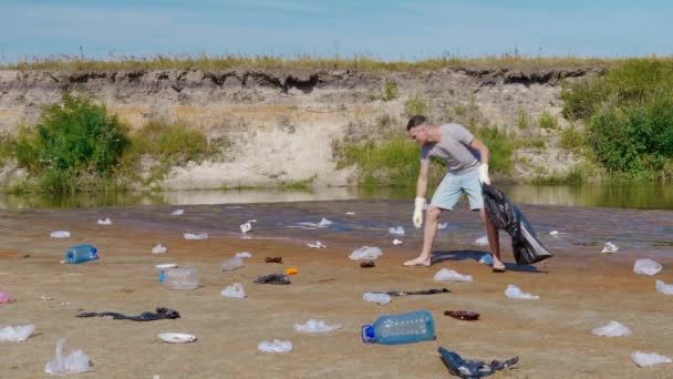 Dühös ember műanyag szemetet gyűjt a szennyezett folyó partján, szemetet dobál. — Stock videók