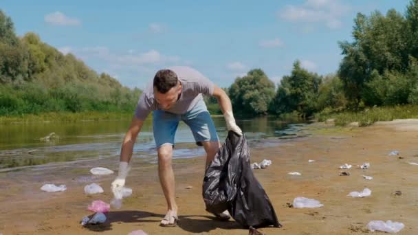 Homem irritado coleta lixo de plástico nas margens do rio poluído, cai lixo — Vídeo de Stock