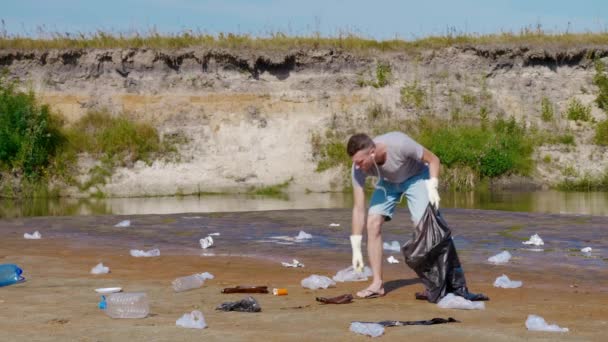 Чоловік збирає пластикові сміття на берегах забрудненої річки і слухає музику — стокове відео