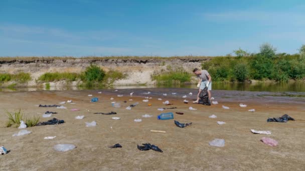 Человек собирает пластиковый мусор на берегу загрязненной реки и слушает музыку — стоковое видео