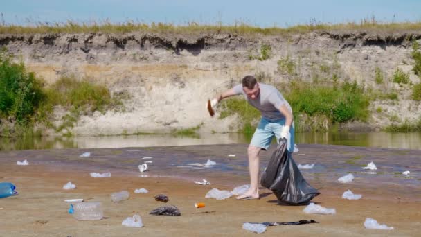 Angry man collects plastic trash on banks of polluted river, then drops garbage — Stock Video