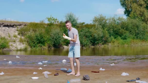 Man picks up plastic trash on the banks of polluted river and shows dislike. — Stock Video