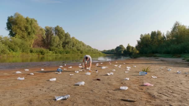Adam kirlenmiş nehrin kıyısından plastik çöpleri topluyor ve sevmediğini gösteriyor.. — Stok video