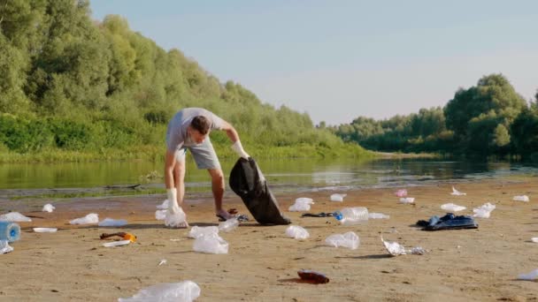 Boze man verzamelt plastic afval op de oevers van vervuilde rivier, en gooit dan afval. — Stockvideo