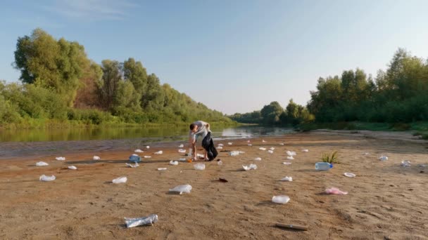 グレーのTシャツとブルーのデニムのショートパンツの男は 乾燥した汚染された川や湖のほとりにプラスチックごみを収集し 親指を表示されます 生態学的大惨事人為的影響だ4K映像 — ストック動画