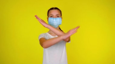 Teen girl in medical mask looks at camera and keeps her arms crossed. Epidemic precautions. Acute Respiratory Virus Infection. Illness cold flu. Stop the virus. Yellow background with copy space. 4K 