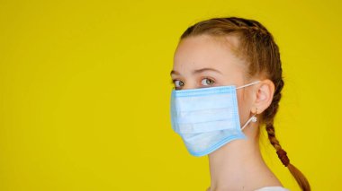 Teen girl in a medical mask looks at camera on yellow background