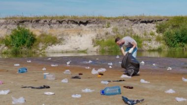 Adam kirlenmiş nehir kıyısında plastik çöp topluyor ve müzik dinliyor.