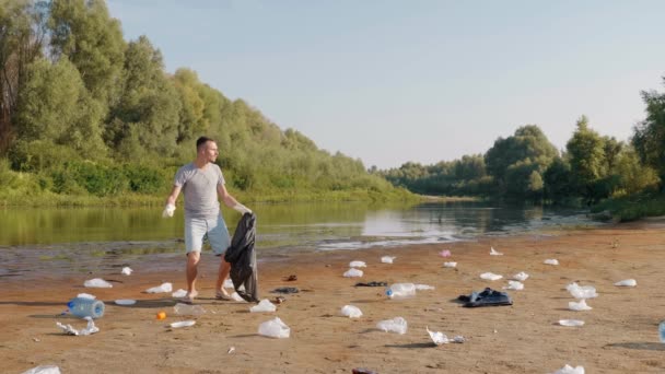 Muž tančí a sbírá plastové odpadky na březích suché a znečištěné řeky — Stock video