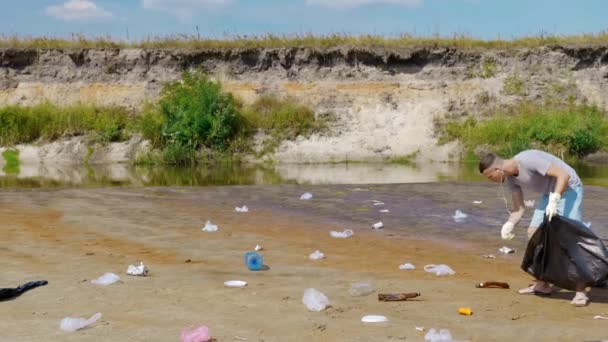 Человек иколлирует пластиковый мусор на берегу загрязненной реки и слушает музыку — стоковое видео