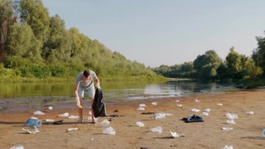 Adam kirlenmiş nehrin kıyısında plastik çöp topluyor ve baş parmağını kaldırıyor.