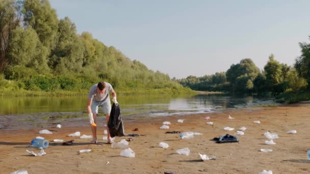 Homem recolhe lixo plástico nas margens do rio poluído e mostra polegares para cima — Vídeo de Stock