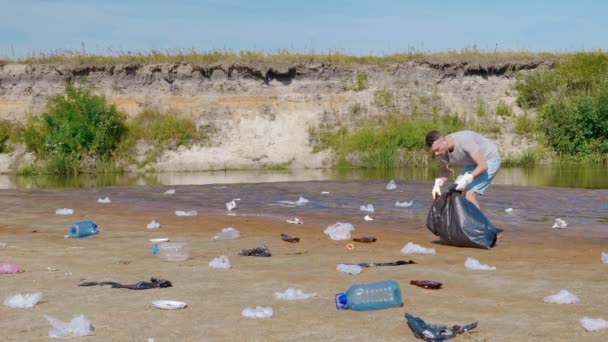 El hombre idancing y recoge la basura plástica en las orillas del río seco y contaminado — Vídeos de Stock