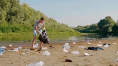 Gri tişörtlü ve mavi kot şortlu bir adam dans ediyor ve kurumuş ve kirlenmiş nehir, göl kıyısında plastik çöp topluyor. Ekolojik felaket. Antropojenik etki. 4k yavaş çekim görüntüsü.