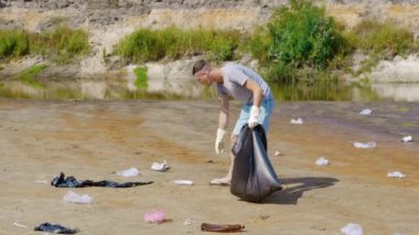 Gri tişörtlü ve kot şortlu bir adam kurumuş ve kirlenmiş bir nehir veya gölün kıyısında plastik çöp topluyor ve kulaklıkla müzik dinliyor. Ekolojik felaket. Antropojenik etki. 4k