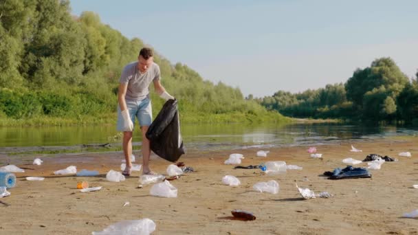Adam kirlenmiş nehrin kıyısında plastik çöp topluyor ve müzik dinliyor. — Stok video