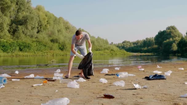 De mens verzamelt plastic afval op de oevers van vervuilde rivier en luistert naar muziek — Stockvideo