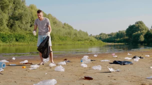 De mens verzamelt plastic afval op de oevers van droge en vervuilde rivier of meer en luistert favoriete lied, meezingen. Ecologische catastrofe. Antropogene invloed. 4k slow motion beelden. — Stockvideo