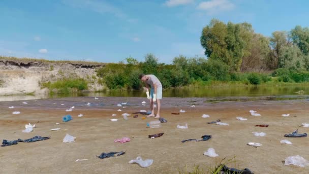 Man in a gray t-shirt and blue denim shorts is picks up plastic trash on the banks of a dry and polluted river or lake and points to it. Ecological catastrophy. Anthropogenic influence. 4K footage. — ストック動画