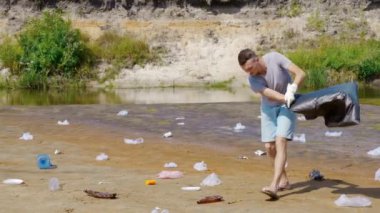 Kızgın adam kirlenmiş nehir kıyısında plastik çöp topluyor sonra da çöp atıyor.