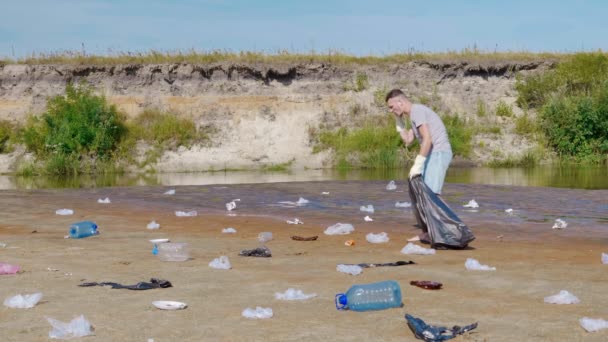 Człowiek zbiera plastikowe śmieci na brzegach zanieczyszczonej rzeki i słucha ulubionej piosenki — Wideo stockowe