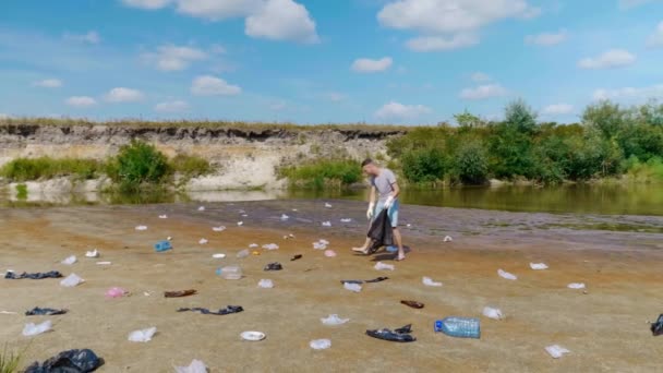 Un hombre pantalones cortos se recoge basura de plástico en las orillas del río contaminado — Vídeos de Stock