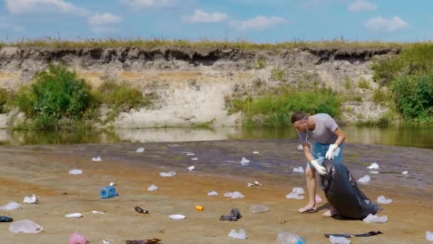 El hombre recoge basura plástica a orillas del río contaminado y escucha música — Vídeos de Stock