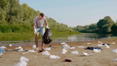 Adam kirlenmiş nehir kıyısında plastik çöpleri topluyor ve baş parmağını kaldırıyor.