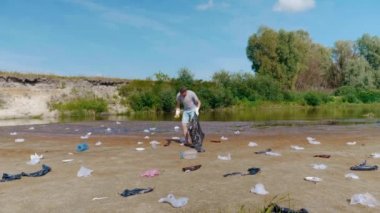 Adam kirlenmiş nehir kıyısında plastik çöpleri topluyor ve baş parmağını kaldırıyor.