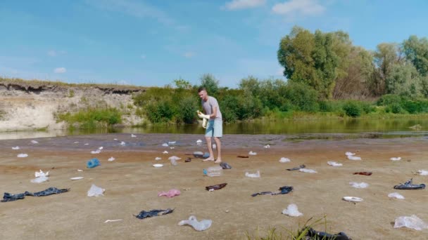 Człowiek zbiera plastikowe śmieci na brzegach zanieczyszczonej rzeki i pokazuje, że nie lubi — Wideo stockowe