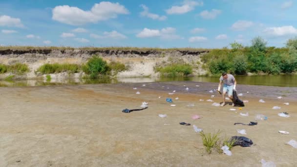 El hombre está bailando y recoge basura plástica en las orillas del río contaminado — Vídeos de Stock