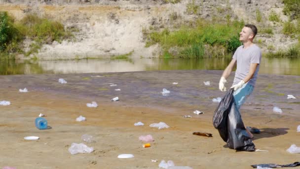 Homem coleta lixo de plástico nas margens do rio poluído e ouve música favorita — Vídeo de Stock