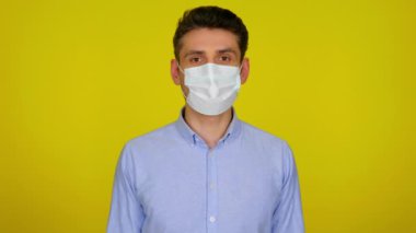 Tıbbi maskeli genç adam sarı arka plandaki kameraya bakıyor.